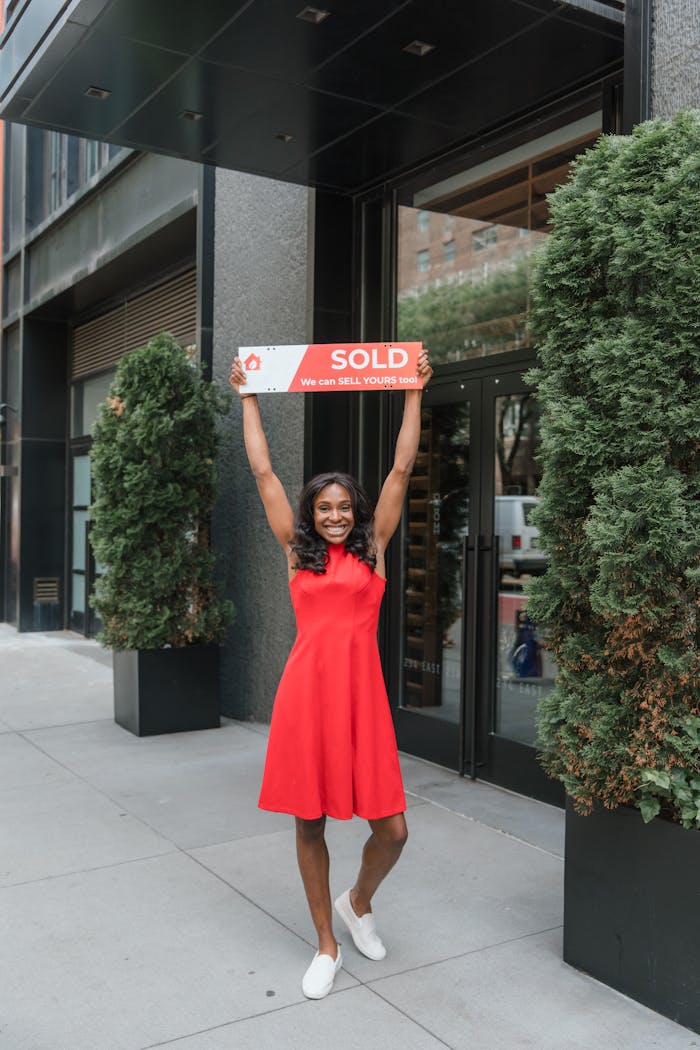 Smiling woman in red dress celebrating real estate sale with a sold sign outdoors.
